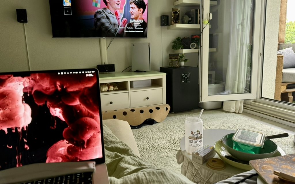 Blick vom Sofa: der Fernseher läuft, davor ein Laptop, auf dem Couchtisch warten benutztes Geschirr und eine Pfanne.