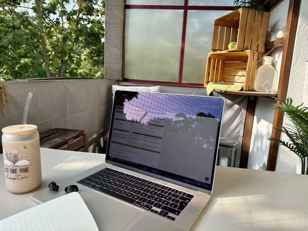 Aufgeklappter Laptop mit Kopfhörern und einem kalten Getränk auf einem sonnigen Balkontisch.