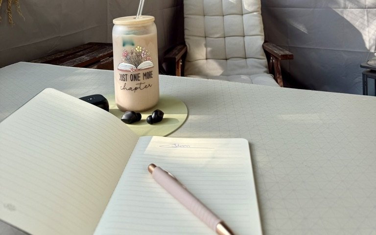 Aufgeschlagenes Notizbuch mit Stift, daneben ein Becher mit Buch-Motiv, auf einem ruhigen Balkontisch im Sonnenlicht.